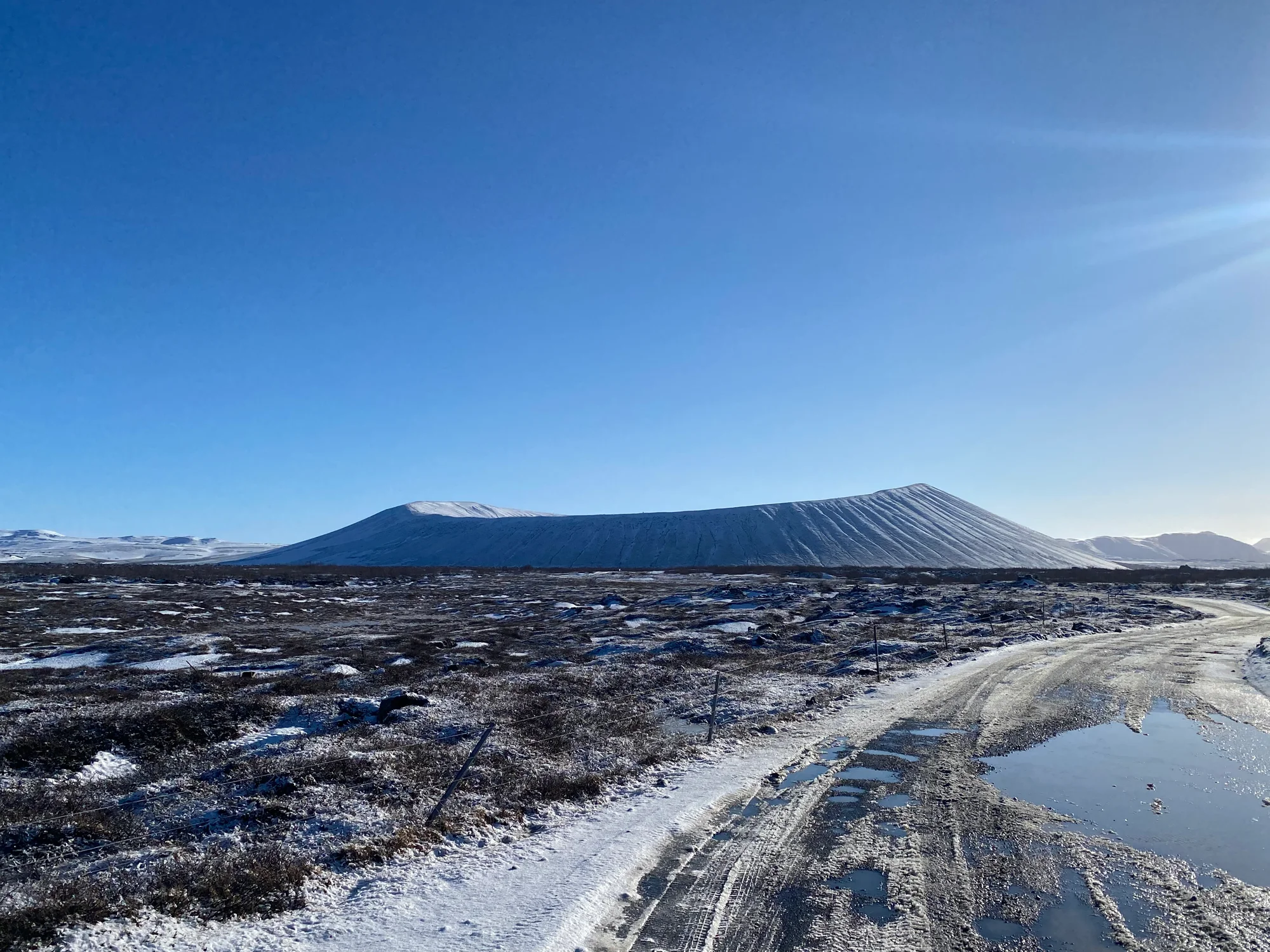 Snow-dusted volcanic terrain beneath a blue sky with a road in the foreground